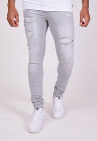 Jeans skinny gris desgastados con múltiples rasgaduras y bordes deshilachados, combinados con zapatillas blancas. Diseño simple, sin patrones o acentos adicionales.