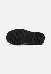 Suela de zapato de goma negra con un patrón de banda de rodadura que presenta ranuras horizontales y verticales para mayor tracción. Nombre de la marca grabado en la suela.