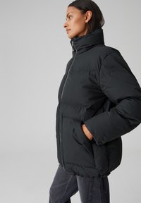 Giacca a vento nera con alto colletto, chiusura con zip e due tasche frontali. Presenta un design trapuntato e un orlo elasticizzato, realizzata in materiale resistente all'acqua.