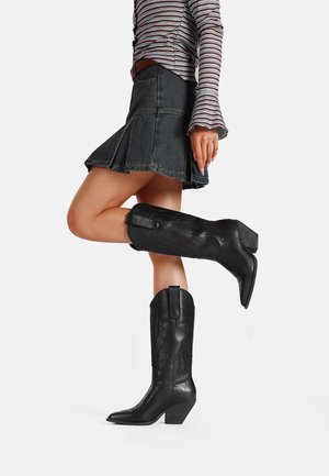 Stivali da cowboy in pelle nera con punta affilata, fiamme testurizzate impresse sui lati e tacco basso a blocco, abbinati a una gonna di denim.