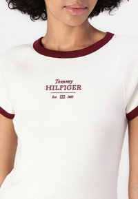 Bela majica s kratkimi rokavi z maroon ovratnikom in manšetami. Ima potiskano logo "Tommy HILFIGER" in "Est. 1985" v maroon barvi. Gladka tekstura.