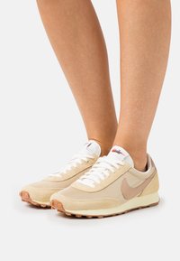 Beige athletic shoes with mesh and suede materials, white laces, and a brown rubber sole. Features a prominent swoosh logo on the side.