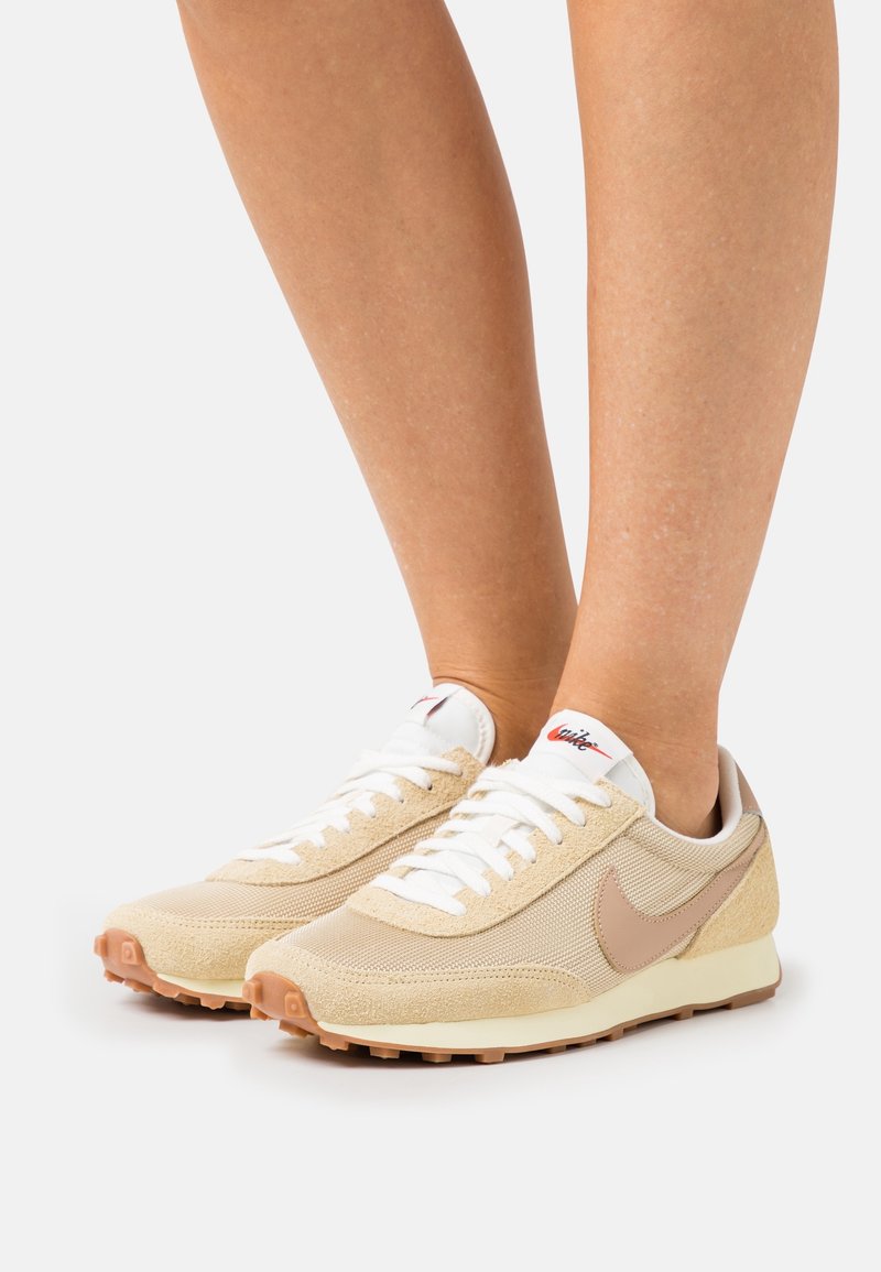 Beige athletic shoes with mesh and suede materials, white laces, and a brown rubber sole. Features a prominent swoosh logo on the side.