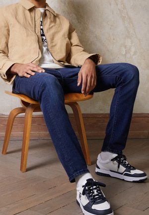 Homme assis sur une chaise en bois, portant une veste beige, un t-shirt blanc à motif graphique, un jean bleu, des baskets blanches et noires, sur un fond de mur clair texturé.