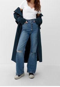 Lång marinblå trenchcoat över en vit ribbad topp, höga blå jeans med slitna hål och ljusa sneakers. Knappar synliga på detaljerna.