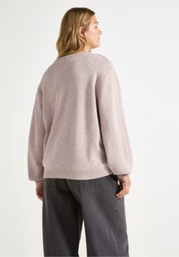 Lys rosa strikket genser med løs passform, rund hals og ribbestrikkede mansjetter, kombinert med mørkegrå vide bukser; teksturert stoff synlig.