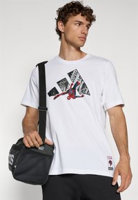 Balta medvilnės marškinėliai su spalvinga Žmogaus voro grafika, juodomis Adidas juostelėmis ir Marvel logotipo pleistru apačioje. Juoda kuprinė su tinklinėmis detalėmis.