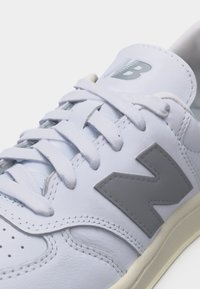 Λευκό δερμάτινο αθλητικό παπούτσι με γκρι λογότυπο "N", υφασμένα κορδόνια και επένδυση στον αστράγαλο σε εκρού σόλα, φωτογραφημένο από κοντινή γωνία.