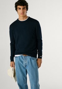 Suéter de cuello redondo en azul marino de tejido suave, combinado con jeans de mezclilla azul claro. Camiseta blanca visible en el escote, llevando una gorra beige.