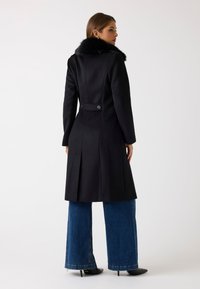 Sort uldfrakke med faux fur-krave, rygslids og knapdetaljer, parret med brede denimjeans og hælede sko.
