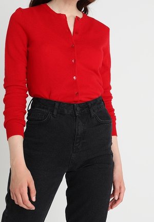Woman wearing a fitted red buttoned cardigan and high-waisted black jeans, standing against a plain white background.