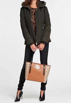 Femme portant une doudoune noire, un haut à imprimé léopard, un pantalon noir, des talons noirs, tenant un sac fourre-tout beige et marron clair avec des poignées noires.