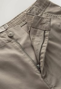 Jasnobeżowe spodnie z częściowo rozpiętym zamkiem błyskawicznym, guzikiem i zapięciem na rzep, ukazujące szczegóły wewnętrznej talii.