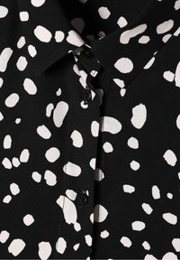 Tissu noir avec des taches blanches irrégulières, doté d'un col plié et de boutons noirs sur le devant.