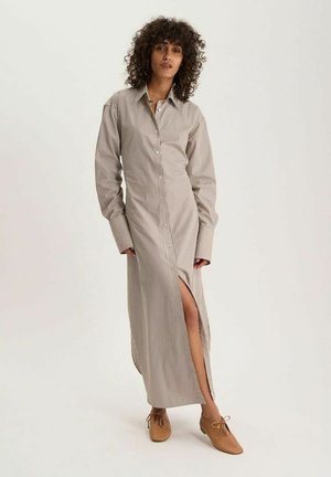 Femme aux cheveux bouclés portant une robe chemise beige à rayures, manches longues, fente à l'avant et chaussures lacées couleur camel, debout sur un fond uni.