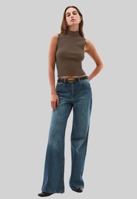 Bruine mouwloze top gecombineerd met lichtblauwe wijde jeans. De jeans zijn voorzien van een riem met een ronde metalen gesp. Gladde textuur overal.