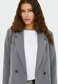 Pelēks žakets ar strukturētu dizainu, diviem priekšējiem pogām un izgrieztām atlokām, uzvilkts virs baltas kreklas. Tekstūras audums ar niansētiem raksta elementiem.