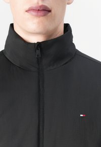Svart hettejakke med høy krage, med glidelås foran og en liten flerfarget logo på venstre side av brystet.