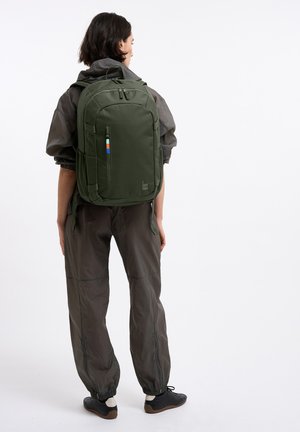 Persona che indossa un abbigliamento scuro e scarpe da ginnastica nere con calzini bianchi, che porta uno zaino verde oliva grande con cerniera frontale e tasche laterali.