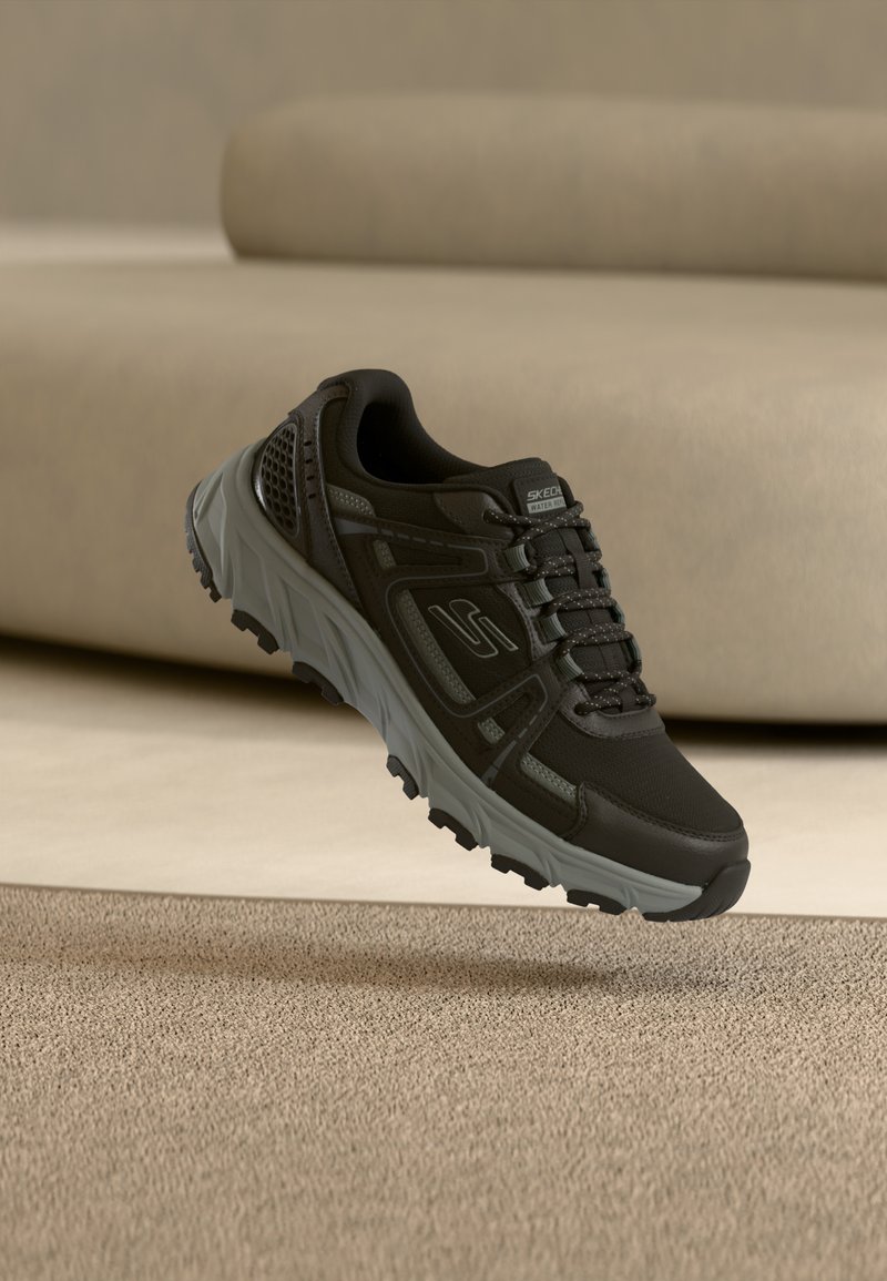 Μαύρο αθλητικό παπούτσι Skechers με γκρι σόλα και αντανακλαστικές λεπτομέρειες, να αιωρείται πάνω από ένα μπεζ χαλί και ουδέτερο φόντο από καναπέ.