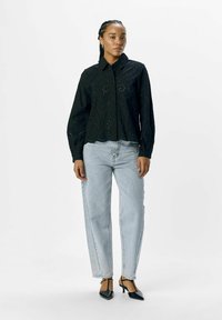 Zwarte, lange mouwen shirt met een gegolfde zoom en oogjesdetail, gecombineerd met lichtblauwe, ontspannen pasvorm jeans en zwarte, spitse hakken.