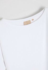 Biała koszulka z szerokim dekoltem, krótkimi rękawami, wykonana z miękkiego materiału. Posiada minimalistyczny design oraz małą metkę marki z tyłu.