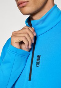 Τεχνητή μπλε ζακέτα με μισό φερμουάρ φτιαγμένη από λείο ύφασμα, διαθέτοντας μαύρο φερμουάρ και λογότυπο «COLMAR» εκτυπωμένο μπροστά.