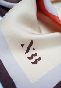 Silk scarf featuring a cream base with geometric patterns in purple and red tones, adorned with dark brown branding elements.