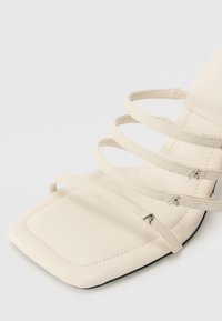 Sandale à talons beige à lanières avec un carré, fabriquée en matériau lisse. Présente plusieurs fines lanières et des accents métalliques subtils.