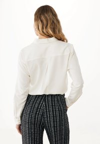 Camicia bianca con maniche lunghe e piega sul retro, abbinata a pantaloni a quadri bianco e nero di tessuto testurizzato, caratterizzati da un design a vita alta.