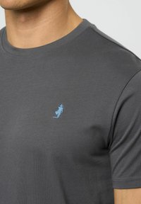 Grå t-skjorte med rund hals, korte ermer og en liten blå brodering på venstre bryst. Myk, jevn tekstur.