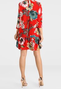Červené květinové šaty s tříčtvrtečními rukávy, volánkovým spodním lemem a klíčovou dírou na zadní straně. Materiál ukazuje velké vícebarevné květinové vzory.