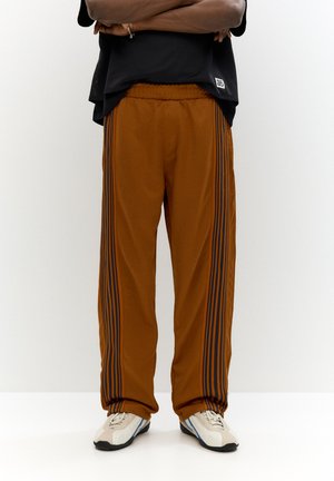 Brown track pants with black side stripes, elastic waistband, and loose fit. Paired with light-colored athletic shoes.