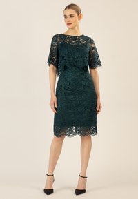 Spitzenkleid in dunklem Petrol mit floralem Muster. Ausgestellte Ärmel und gezackter Saum. Kombiniert mit schwarzen spitzen Absätzen.