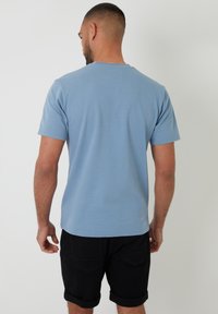 Světle modré tričko s krátkým rukávem, kulatým výstřihem a texturovanou látkou, nošené s černými šortkami, z pohledu zezadu.