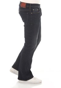 Jean en denim bleu foncé avec une taille ajustée, une ouverture de jambe évasée et des coutures visibles. Présente une étiquette de marque en cuir marron à l'arrière.