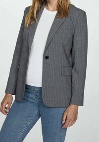 Blazer gris a rayas con un ajuste estructurado. Cuenta con un cierre de un solo botón, solapas de muesca y bolsillos frontales. Llevado sobre una camisa blanca.