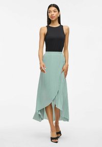 Black sleeveless top paired with a flowing, pleated mint-green skirt featuring a high-low hem and black open-toe heeled shoes.