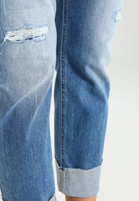 Jeans de denim azul claro con características desgastadas, con dobladillos y puños deshilachados. La tela muestra un leve desvanecimiento y un diseño de corte holgado.