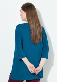 Túnica color teal con mangas tres cuartos, con un escote redondeado y aberturas laterales, combinada con pantalones color burdeos. Tejido suave, con detalles mínimos.