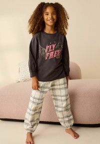 Dunkelgraues Langarmshirt mit dem Aufdruck "FLY FREE", kombiniert mit cremefarbenen Karohosen. Im Hintergrund steht eine rosa Couch mit einer texturierten Oberfläche.
