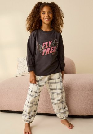 Ciemnoszara koszula z długim rękawem z grafiką "FLY FREE", zestawiona z kremowymi spodniami w kratę. Kanapa w tle jest różowa z teksturowaną powierzchnią.