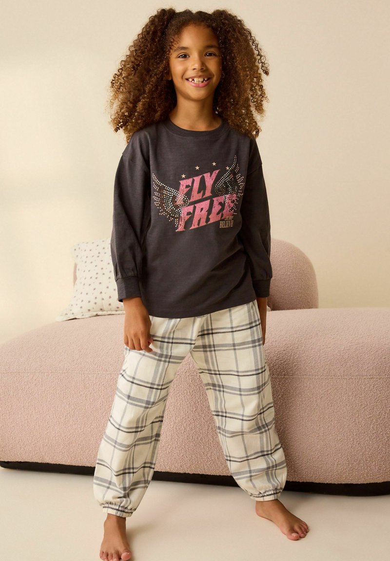 Cămașă cu mâneci lungi de culoare gri închis, cu grafică "FLY FREE", asortată cu pantaloni crem cu carouri. Canapeaua din fundal este roz, cu o suprafață texturată.