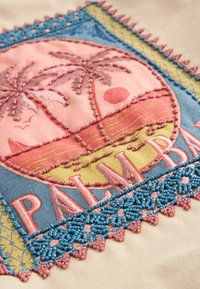Broderie d'un coucher de soleil tropical avec des palmiers, un voilier, des oiseaux et le mot "PALM BEACH" entouré de motifs décoratifs en perles bleues et roses.
