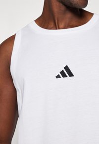 Bela brezrokavna športna majica iz bombaža, ki ima črn izšit Adidas logo na prsih. Gladka tekstura in okroglo izrez.