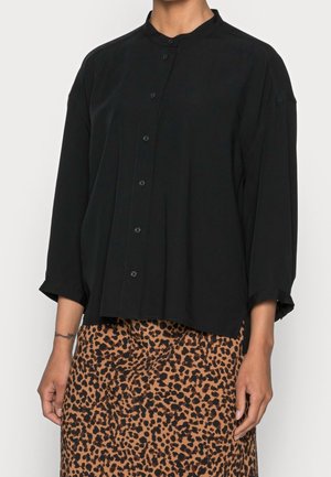 Vrouw draagt een zwart overhemd met lange mouwen en een mandarijnkraag, gecombineerd met een bruine rok met luipaardprint.