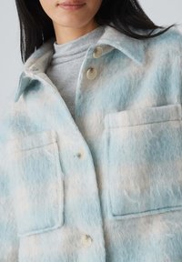 Veste en laine duveteuse bleu clair avec des accents beiges, dotée d'une fermeture à boutons, de deux grandes poches plaquées et d'un col. Texture douce.