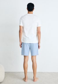 Witte katoenen T-shirt en lichtblauwe shorts, eenvoudig ontwerp, gedragen door een persoon die blootsvoets staat, gezien van achteren tegen een neutrale achtergrond.