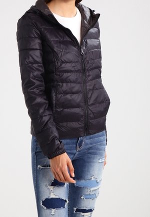 Personne portant une veste matelassée noire zippée avec capuche et un jean bleu déchiré, debout devant un fond clair uni.