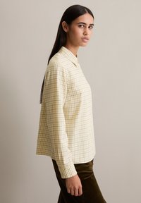 Yellow plaid shirt with a collar and long sleeves. The fabric features a grid pattern with contrasting dark lines. Simple and structured design.